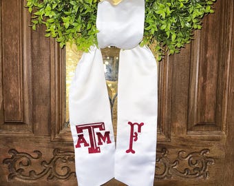 May include: A white graduation stole with maroon embroidered letters "A T M" and "F". The stole is hanging on a wooden door with a green wreath in the background. The letters are in a collegiate style.