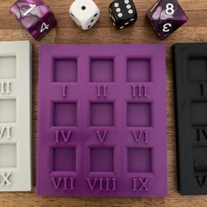 May include: A selection of dice and dice moulds. The dice are white, black, and purple. The moulds are white, purple, and black, each with Roman numerals. The dice are for tabletop games.