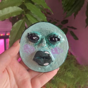 Può includere: Un pezzo decorativo rotondo, color verde acqua, con un volto scolpito. Il volto ha occhi scuri, ciglia nere e labbra verde scuro con accenti bianchi. Le guance sono dipinte di un colore viola chiaro.