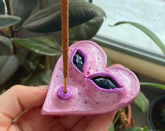 Könnte beinhalten: Ein rosa herzförmiger Räucherstäbchenhalter mit zwei schwarzen, augenförmigen Öffnungen. Der Halter hat eine strukturierte Oberfläche und einen kleinen lila Kreis am Boden. Ein Räucherstäbchen wird in den Halter gesteckt. Der Halter wird in einer Hand gehalten.