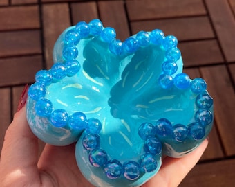Könnte beinhalten: Hellblaue, blütenförmige Schmuckschale mit Perlenrand. Die Schale wird in einer Hand gehalten und hat ein glänzendes Finish. Die Perlen sind von transluzenter blauer Farbe.
