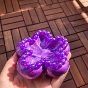 K&ouml;nnte beinhalten: Eine lila, bl&uuml;tenf&ouml;rmige Schmuckschale mit gl&auml;nzendem Finish. Die Schale ist mit kleinen, irisierenden lila Perlen am Rand verziert. Die Schale wird in einer Hand gehalten.