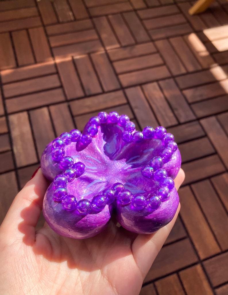 K&ouml;nnte beinhalten: Eine lila, bl&uuml;tenf&ouml;rmige Schmuckschale mit gl&auml;nzendem Finish. Die Schale ist mit kleinen, irisierenden lila Perlen am Rand verziert. Die Schale wird in einer Hand gehalten.