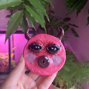 May include: A pink, round, devil-faced sculpture with dark brown horns, a dark brown snout, and large black eyes. The sculpture has pink cheeks and is held in a hand. The background includes green plants and a pinkish light.