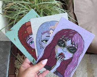 Könnte beinhalten: Drei quadratische Kunstdrucke mit Illustrationen stilisierter weiblicher Gesichter in kräftigen Farben. Die Drucke werden in einer Hand gehalten, mit einer Pflanze im Hintergrund. Die Illustrationen haben einen einzigartigen, künstlerischen Stil.