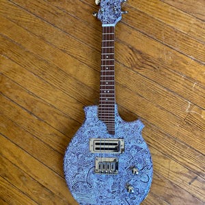 May include: A custom-made electric guitar with a unique, patterned design. The body and headstock feature a blue and white doodle pattern. The guitar has a brown neck, gold hardware, and two control knobs. The instrument is displayed on a wooden floor.