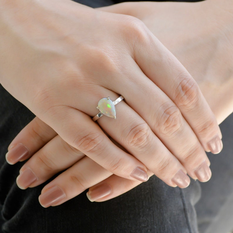May include: A pearlescent teardrop-shaped opal ring set in a silver band. The ring is on a person's finger.