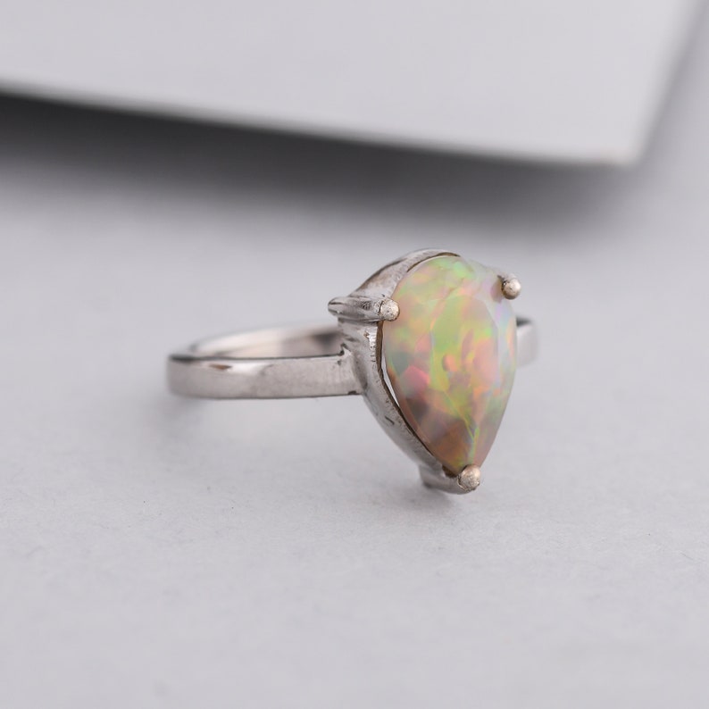 May include: A silver ring with a pear-shaped opal gemstone. The opal has a rainbow of colors, including green, yellow, and pink.