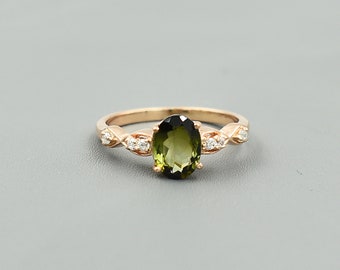 May include: Rose gold ring with an oval green gemstone. The band has small, clear accent stones and a decorative design on either side of the main stone. The ring is set against a grey background.