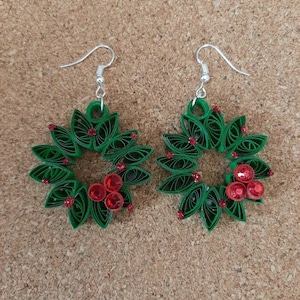 May include: A pair of green paper quilled earrings shaped like Christmas wreaths with red glitter embellishments.