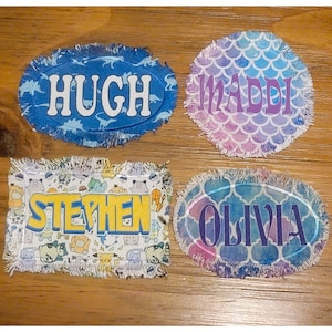 May include: Four personalized fabric patches with frayed edges. The patches feature the names "Hugh", "Maddi", "Stephen", and "Olivia" in different fonts and colors. The patches are decorated with various designs, including dinosaurs, mermaids, and cartoon characters.