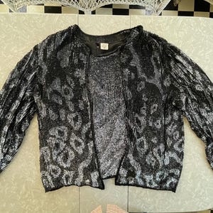 May include: A black and silver sequined jacket with a long sleeve and a round neckline. The jacket has a subtle pattern of small, silver sequins.