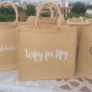 May include: A set of brown burlap tote bags with white lettering. The bags have different phrases printed on them, including "Bride" and "Wifey for Life".