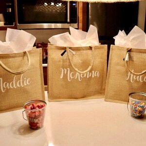 May include: Three brown burlap tote bags with white handles and white personalized names. The names are Maddie, McKenna, and Haley. The bags are sitting on a white surface with small containers of candy in front of them.
