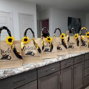 May include: Six brown burlap tote bags with black and yellow trim and a sunflower design. Each bag has a different name printed on it: Nicole, Loletta, Audrio, Tra, Trina, and a. The bags have the text "Women Sip & Sail 2024" printed on them.