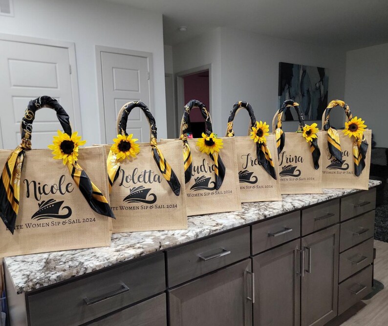 May include: Six brown burlap tote bags with black and yellow trim and a sunflower design. Each bag has a different name printed on it: Nicole, Loletta, Audrio, Tra, Trina, and a. The bags have the text "Women Sip & Sail 2024" printed on them.