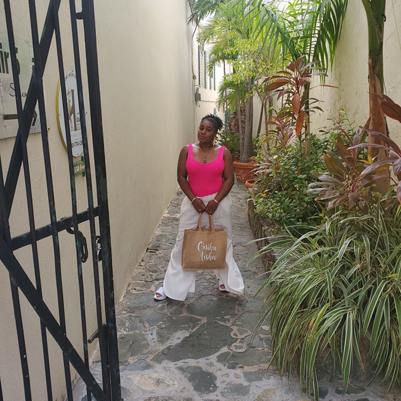 May include: A woman wearing a pink tank top and white pants is standing in a narrow alleyway. She is holding a brown tote bag with the words "Onko Asha" printed on it.