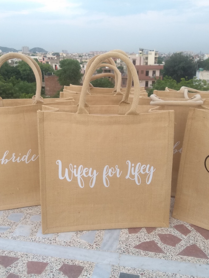 May include: A set of brown burlap tote bags with white lettering. The bags have different phrases printed on them, including "Bride" and "Wifey for Life".