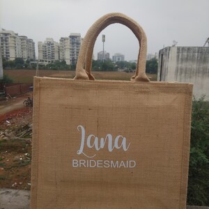 May include: A brown burlap tote bag with white handles and the text "Lana Bridesmaid" printed on the front.