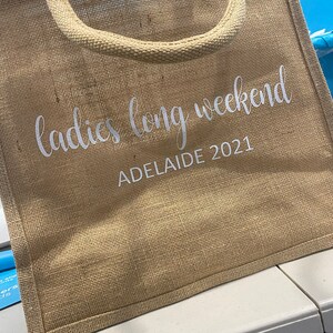 May include: A brown burlap tote bag with white text that reads "ladies long weekend ADELAIDE 2021".