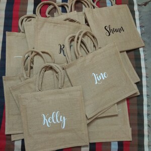 May include: A stack of brown burlap tote bags with white and black lettering. The bags are personalized with the names "Kelly", "Line", "Shawn", and "R".