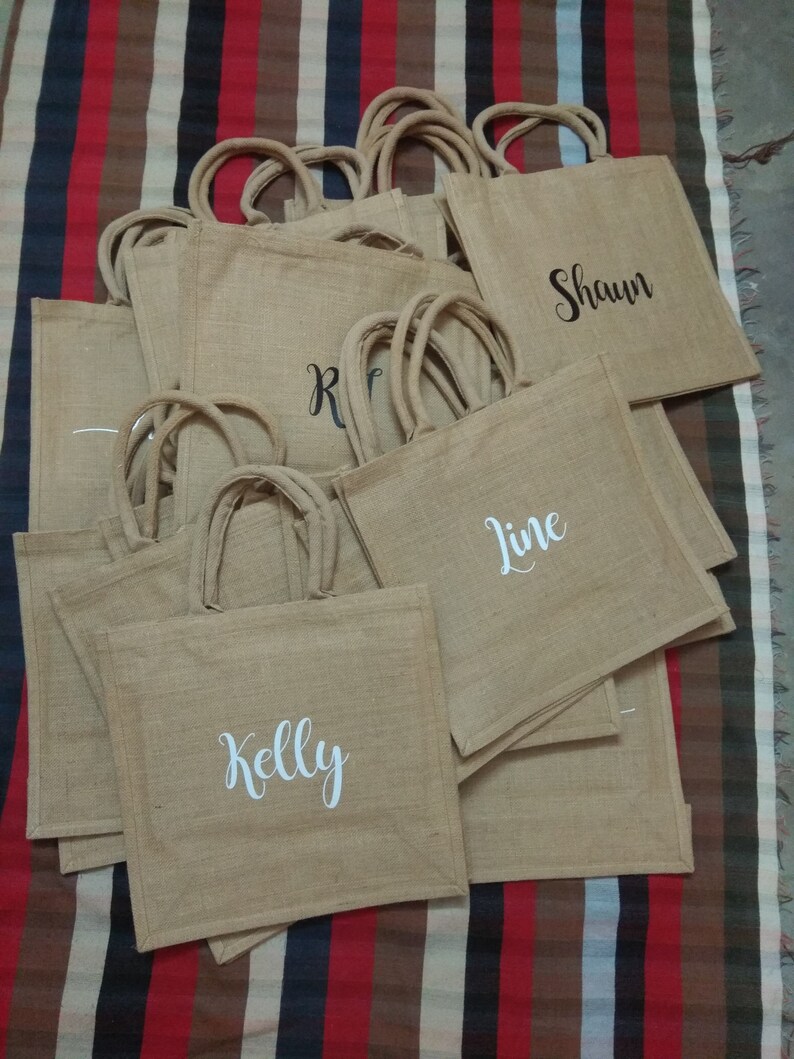 May include: A stack of brown burlap tote bags with white and black lettering. The bags are personalized with the names "Kelly", "Line", "Shawn", and "R".