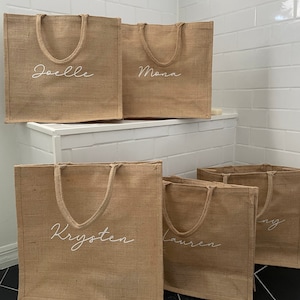 May include: Five brown burlap tote bags with white embroidered names: Joelle, Mona, Krysten, Lauren, and Amy.