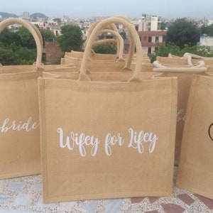 May include: A group of brown burlap tote bags with white lettering. The bags have different phrases printed on them, including "Bride" and "Wifey for Life".