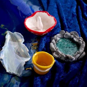 May include: Four small ceramic dishes in different shapes and colours. A white dish with a red rim, a blue dish with a wavy edge, a yellow dish with a white rim, and a grey dish with a green centre.