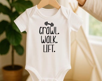 May include: White baby onesie with short sleeves, hanging on a wooden hanger. The onesie features the text "Crawl. Walk. Lift." in black, with a dumbbell graphic above the word "crawl". The onesie is made of soft material and has snap closures.