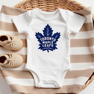 Peut inclure: Body bébé blanc avec le logo bleu marine des Maple Leafs de Toronto. Le logo est une feuille d'érable stylisée avec l'inscription "TORONTO MAPLE LEAFS". Le body est posé sur une couverture rayée avec un panier en osier et deux paires de chaussures de bébé beiges.