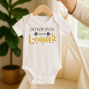 Puede incluir: Body de bebé blanco con el texto "DO YOU EVEN Leviosa?" en letras negras y doradas. El diseño incluye un gráfico de barra y detalles de estrellas. El body está colgado de una percha de madera.