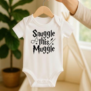 Puede incluir: Body de beb&eacute; blanco con el texto negro "Snuggle this Muggle". El dise&ntilde;o incluye gafas, estrellas y una varita m&aacute;gica. El body es de manga corta y tiene tres corchetes en la parte inferior.