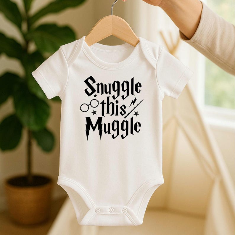 Puede incluir: Body de beb&eacute; blanco con el texto negro "Snuggle this Muggle". El dise&ntilde;o incluye gafas, estrellas y una varita m&aacute;gica. El body es de manga corta y tiene tres corchetes en la parte inferior.