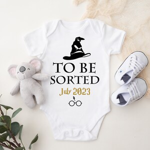 May include: White baby onesie with a black Sorting Hat graphic and the text "TO BE SORTED July 2023" in black and gold. Includes a pair of white baby shoes and a gray koala plush toy.