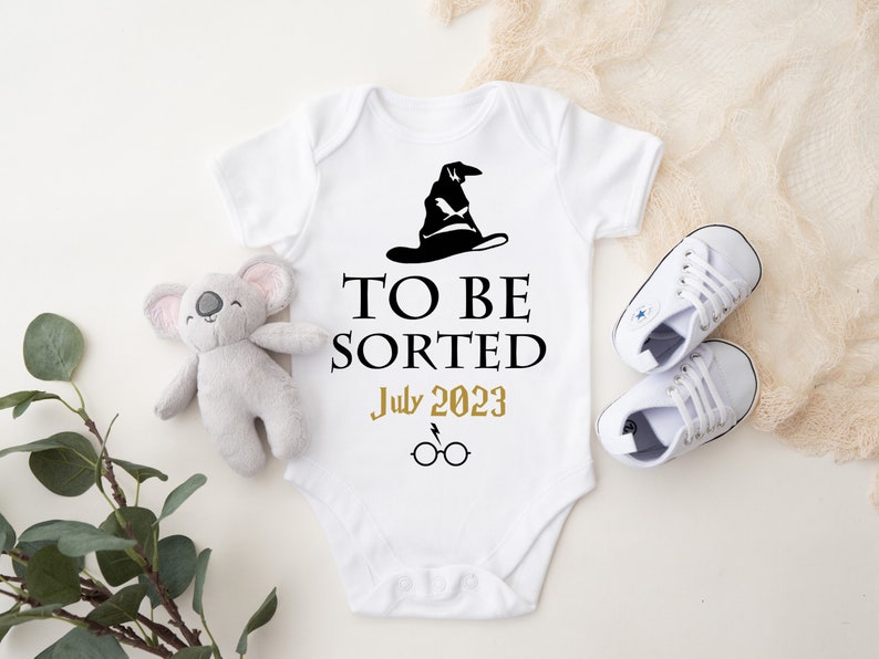 May include: White baby onesie with a black Sorting Hat graphic and the text "TO BE SORTED July 2023" in black and gold. Includes a pair of white baby shoes and a gray koala plush toy.
