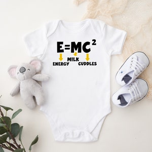 Op de afbeelding: Witte babybody met de tekst "E=MC²" en de woorden "Milk, Energy, Cuddles" eronder. Een grijs koala knuffel en een paar witte babyschoenen liggen naast de body. De body is een babykledingstuk.