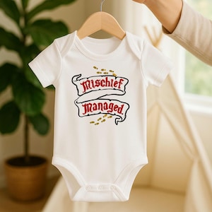 Peut inclure: Body bébé blanc avec l'inscription "Mischief Managed" dans un motif de bannière. Le texte est rouge et noir avec des empreintes de pas dorées. Le body est suspendu à un cintre en bois.
