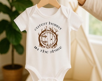May include: A white short-sleeved baby bodysuit featuring a brown graphic of a round door set within tree branches. The text "CUTEST HOBBIT IN THE SHIRE" is printed in a dark brown font, arching above and below the central circular door illustration.