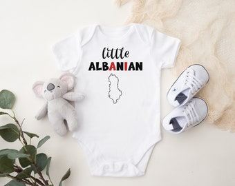 May include: White baby onesie with the text "little ALBANIAN" in black and red, and a map outline of Albania. A gray koala plush toy and a pair of white baby sneakers are next to the onesie.