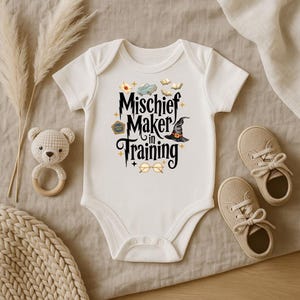 May include: A white short-sleeved baby bodysuit featuring the black text "Mischief Maker in Training" surrounded by small illustrations of a blue car, open books, a witch hat, a pentagonal badge, and round glasses with gold star accents.