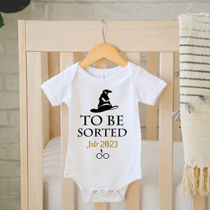 May include: White baby onesie with a black sorting hat graphic and the text "TO BE SORTED July 2023" in black and gold. The onesie is hanging on a wooden hanger.