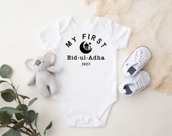 May include: White baby onesie with the text "MY FIRST Eid-ul-Adha 2023" in black. The design includes a crescent moon and a star. A gray koala plush toy and a pair of white baby shoes are next to the onesie.