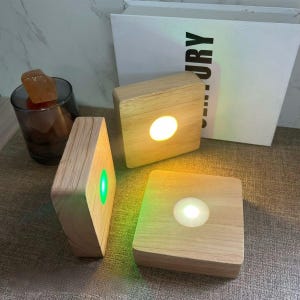 May include: Three wooden display bases with built-in LED lights. Each base is square or trapezoidal, with a circular light in the centre. The lights glow in white, green, and yellow. A glass of salt crystals is in the background.