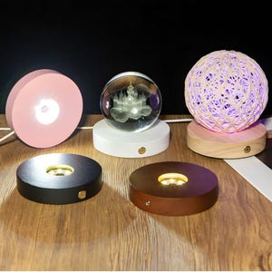 Könnte beinhalten: Eine Sammlung beleuchteter Display-Sockel in verschiedenen Farben und Designs. Enthält einen rosa kreisförmigen Sockel, einen weißen Sockel mit einer Glaskugel mit Schlossdesign und einen Holzsockel mit einer gewebten Kugel. Auch schwarze und braune Sockel sind abgebildet.