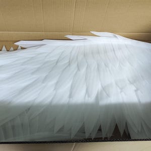 May include: A pair of large, white, feathered-style wings constructed from layered, pointed foam or plastic segments. The wings are positioned horizontally within a brown cardboard shipping box, displaying a detailed, overlapping wing structure.