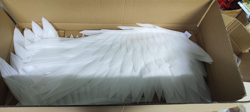 May include: A pair of large, white, feathered-style wings constructed from layered, pointed foam or plastic segments. The wings are positioned horizontally within a brown cardboard shipping box, displaying a detailed, overlapping wing structure.