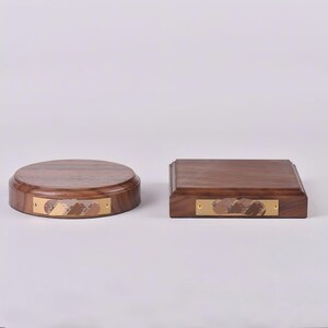 May include: Two wooden display bases. One is circular, the other is square. Both are made of dark wood and feature a rectangular brass plate with a decorative design. The bases are suitable for displaying various items.