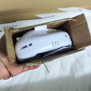 May include: A white plastic electronic device with a smooth, curved top surface and a small rectangular protrusion, partially contained within an open brown cardboard box. The device features visible side vents and a small grey component underneath.