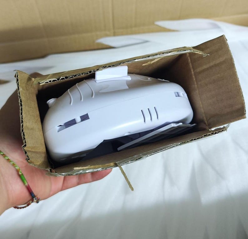 May include: A white plastic electronic device with a smooth, curved top surface and a small rectangular protrusion, partially contained within an open brown cardboard box. The device features visible side vents and a small grey component underneath.